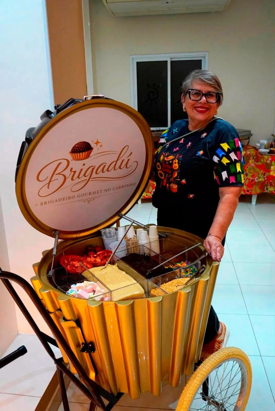 Promotora do Brigadú ao lado do carrinho dourado de brigadeiro gourmet em uma festa junina